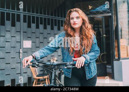 Rot gewellt lange Haare kaukasischen Teenager-Mädchen auf der Stadt Straße zu Fuß mit Fahrrad Mode Porträt. Natürliche Menschen Schönheit urbanes Leben Konzept Bild. Stockfoto