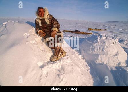 Inuit älterer Mann, Mitte 60, in traditioneller Karibu-Kleidung gekleidet, auf einem traditionellen Iglu. Tatsächliche Iglus werden nicht mit Blöcken zurückgelassen, die angezeigt werden. Wenn die Blöcke an Ort und Stelle sind, ist das Iglu mit Schnee bedeckt, um weitere Isolierung zu erhalten. Ein traditioneller Schlitten (Komatik) ist im Hintergrund zu sehen. Stockfoto