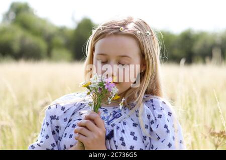 Ein junges hübsches Hippie-Mädchen mit blonden Haaren hält im Sommer in einem Weidefeld eine schicke wilde Blume im Sonnenschein. Stockfoto