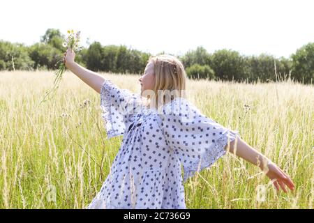 Ein junges hübsches Hippie-Mädchen mit Gänseblümchen-Kette in blonden Haaren hält im Sommer eine schicke wilde Blume mit ausgestreckten Armen auf einem Weidefeld Stockfoto