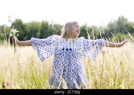 Ein junges hübsches Hippie-Mädchen mit Gänseblümchen-Kette in blonden Haaren hält im Sommer eine schicke wilde Blume mit ausgestreckten Armen auf einem Weidefeld. Stockfoto