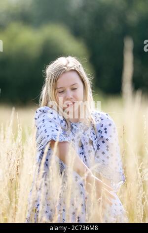 Ein junges hübsches Hippie-Mädchen mit einer Gänseblümchen-Kette in ihren blonden Haaren geht im langen Gras im Sonnenschein auf einem Weidefeld im Sommer. Stockfoto