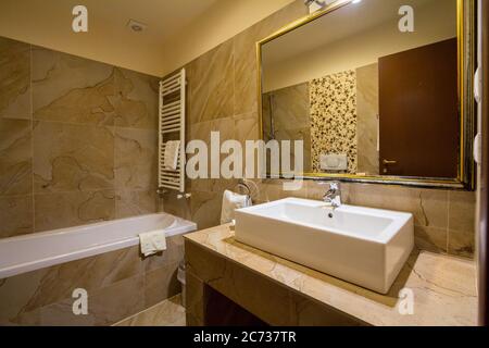 Interieur eines mittelklassigen Hotelbadezimmers mit seiner typischen Badewanne, einem Waschbecken mit Wasserhahn, etwas Toilette, sowie frischen Türmen und einem großen Spiegel, mit einem Stockfoto