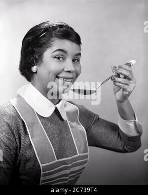 1950S 1960S AFROAMERIKANISCHE FRAU HÄLT LÖFFEL VERKOSTUNG TRAGEN SCHÜRZE HAUSFRAU KOCHEN BLICK AUF KAMERA - N153 HAR001 HARS B&W BLICKKONTAKT ZIELE ERFOLG HAUSFRAU GLÜCK HAUSFRAUEN AFRO-AMERIKANER AFRO-AMERIKANISCHE SCHWARZE ETHNIE STOLZ GESCHMACK VERKOSTUNG HAUSFRAUEN PROBE STILVOLLE MITTE ERWACHSENE FRAU PROBE SCHWARZ UND WEISS LEBENSMITTEL VORBEREITUNG HAR001 ALTMODISCH AFRIKANISCH AMERIKANER Stockfoto