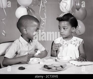 1960S ZWEI AFROAMERIKANISCHE KINDER JUNGE MÄDCHEN BRUDER SCHWESTER EINANDER ANGUCKEN ESSEN EIS COOKIES GEBURTSTAG PARTY - N2081 HAR001 HARS GESICHTS KOMMUNIKATION COMIC FREUDE LIFESTYLE FRAUEN BRÜDER HEIM LEBEN KOPIEREN RAUM FREUNDSCHAFT HALBE LÄNGE MÄNNER COOKIES GESCHWISTER SCHWESTERN AUSDRÜCKE B&W RUNZELIG HUMORVOLL FRÖHLICH AFROAMERIKANISCH AFROAMERIKANISCH GENIESSEN KOMISCH BEI GESCHWISTER LÄCHELT KOMÖDIE FRÖHLICH EIS WACHSTUM JUGENDLICHE ZWEISAMKEIT SCHWARZ UND WEISS EINANDER HAR001 ALTMODISCHE AFROAMERIKANER Stockfoto