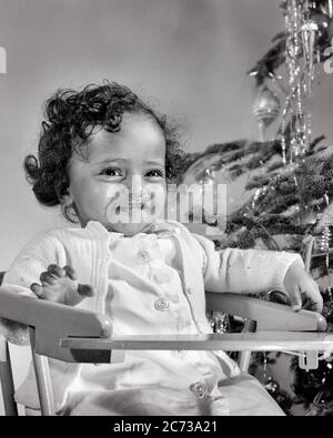 1940ER-JAHRE 1940ER-JAHRE LÄCHELNDES AFROAMERIKANISCHES BABY MÄDCHEN IM HOCHSTUHL NEBEN WEIHNACHTSBAUM - N472 HAR001 HARS WEIBCHEN STUDIO SCHUSS GESUNDHEIT HEIM LEBEN KOPIEREN RAUM HALBE LÄNGE B&W GLÜCK FRÖHLICH AFRO-AMERIKANER FRÖHLICHE AFROAMERIKANISCHE ORNAMENTE SCHWARZE ETHNIE LÄCHELT DEZEMBER 25 NEBEN FRÖHLICHEM WACHSTUM FRÖHLICHE JUGENDLICHE BABY MÄDCHEN SCHWARZ UND WEISS HAR001 ALT ALTMODISCHE AFROAMERIKANER Stockfoto