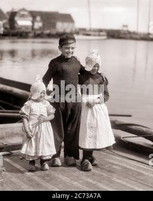 1920ER JAHRE HOLLÄNDISCHER JUNGE STEHEND MIT DEN ARMEN UM ZWEI SCHWESTERN ALLE TRAGEN HOLZSCHUHE UND TRADITIONELLE LOKALE KLEIDUNG VOLLENDAM HOLLAND - R4033 HAR001 HARS JUGENDLICHER STIL TEAMARBEIT FREUDE FREUDE LIFESTYLE ZUFRIEDENHEIT GESCHICHTE FRAUEN BRÜDER GESUNDHEIT HEIM LEBEN KOPIEREN RAUM FREUNDSCHAFT GANZLÄNGE TRADITIONELL MÄNNER GESCHWISTER VERTRAUEN HOLLÄNDISCHE SCHWESTERN EUROPA SCHÜRZEN B&W GLÜCK FRÖHLICH EUROPÄISCH SEINE UND STOLZ LOKALEN GESCHWISTER LÄCHELT HOLLAND FRÖHLICH STYLISH CLOG KOOPERATION JUGENDLICHE PRE-TEEN PRE-TEEN JUNGE DIE NIEDERLANDE ZWEISAMKEIT SCHWARZ UND WEISS KAUKASISCHEN ETHNIZITÄT HAR001 NIEDERLANDE ALTMODISCH Stockfoto