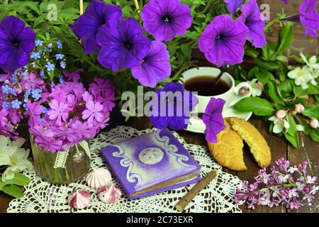 Dekoriertes Buch mit Petunia-Bündel und einer Tasse Tee auf einem Holztisch. Esoterischer, gotischer und okkulter Hintergrund mit magischen Objekten, mystischer und märchenhafter Kon Stockfoto