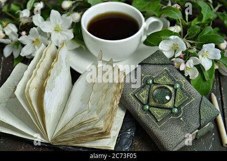 Tagebuchbuch mit einer Tasse Tee und blühenden Apfelbaumbästen auf dem Tisch öffnen. Esoterischer, gotischer und okkulter Hintergrund mit magischen Objekten, Mystiker und Fee Stockfoto