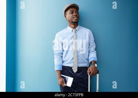 Afrikanischer junger Mann, der an der blauen Wand lehnt, steht er mit Koffer und Tickets und er wird auf Reisen gehen Stockfoto
