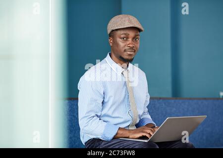 Portrait eines afrikanischen Geschäftsmannes, der die Kamera anschaut, während er mit einem Laptop auf den Knien im Büro sitzt und daran arbeitet Stockfoto