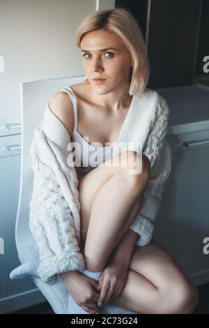 Charmante Dame mit blonden Haaren und Strickpullover sitzt auf dem Stuhl in der Küche und schaut in die Kamera Stockfoto