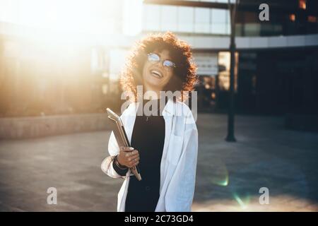 Lockige Geschäftsfrau trägt Brillen und lächelt an der Kamera posiert an einem sonnigen Tag mit einem Laptop Stockfoto