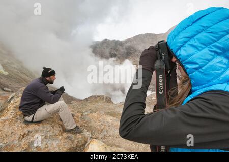 Junge Frau fotografiert Mann in Krater aktiven Vulkan umgeben von Rauch heißen Quellen, Eruption Fumarolen, Gas-Dampf vulkanischen Aktivität Stockfoto