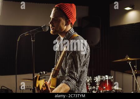 Junge kaukasische wunderschönen Musiker spielen E-Gitarre und singen auf Mikrofon im Aufnahmestudio. rockstar führen Musik. Mann bereitet, üben b Stockfoto