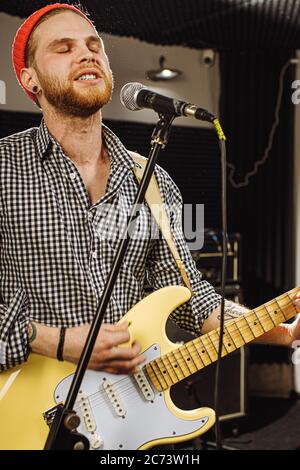 Junge kaukasische Gitarrist gleichzeitig Gitarre spielen und singen ein Lied im Aufnahmestudio. Schöner Kerl in Red hat emotional singen Song, scharf auf mu Stockfoto