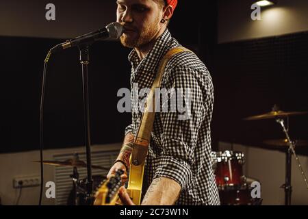 Junge kaukasische wunderschönen Musiker spielen E-Gitarre und singen auf Mikrofon im Aufnahmestudio. rockstar führen Musik. Mann bereitet, üben b Stockfoto