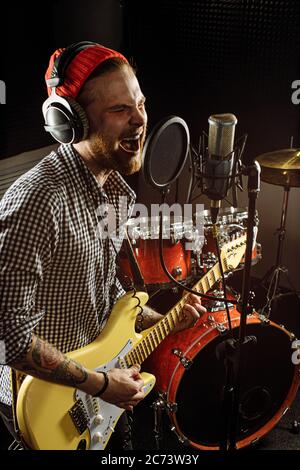 Junge kaukasische wunderschönen Musiker spielen E-Gitarre und singen auf Mikrofon im Aufnahmestudio. rockstar führen Musik. Mann bereitet, üben b Stockfoto