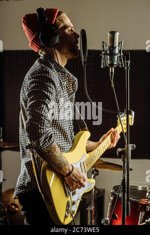 Junge kaukasische Gitarrist gleichzeitig Gitarre spielen und singen ein Lied im Aufnahmestudio. Schöner Kerl in Red hat emotional singen Song, scharf auf mu Stockfoto