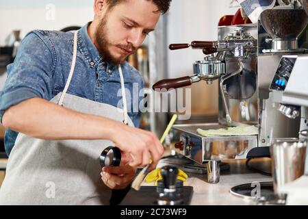Junger bärtiger Mann steht an der Theke, fegt vom Tisch, schaut mit ernstem Ausdruck beiseite, arbeitet hart in der Nähe Kaffeemühle, verwendet Pinsel, Kaffee Stockfoto