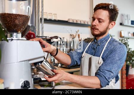 Beschäftigt, seriöse Mann mit dicken Bart in ordentlich Schürze, Sand in der Nähe von Kaffeemaschine, drückt Taste, hält Metall-Kaffeekanne, bereitet Cappuccino für freundliche gue Stockfoto
