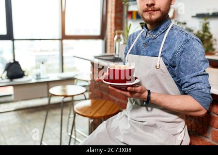 Geschickter talentierter Barkeeper mit dickem Bart trägt weiße Schürze, sitzt auf Holzstuhl, hält leere Kaffeetasse, Small Business-Konzept, Indoor-Shot Stockfoto