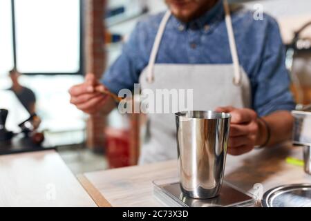 Nahaufnahme von Metall glänzenden Kaffeekanne auf Skala stehen, verschwommener Hintergrund des Mannes hält Silberlöffel mit gemahlenem Kaffee, Barista Messung Kaffee fo Stockfoto