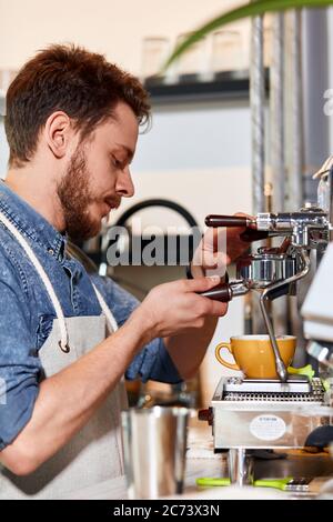 Professioneller Barista, trägt Jeanshemd und gepflegte Schürze, steht an der Kaffeemaschine, hält Temperament, macht Kaffee, schaut weg, konzentriert sich auf Pooring Fres Stockfoto