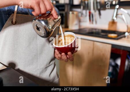 Hände von geschickten jungen Barkeeper Gießen gedämpfte Milch aus Metall Krug, Herstellung von heißen Latte, halten rote große Tasse, Kaffee-Konzept Stockfoto