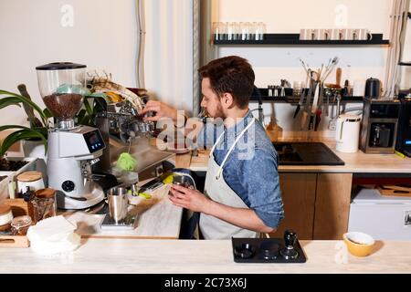 Lächelnder dlad Mann trägt Jeans-Hemd und gepflegte Schürze, steht in der Nähe von professionellen Kaffeemaschine an der Theke, hält Griff, lässt Luft aus, macht frische gro Stockfoto