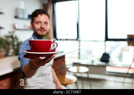 Foto von herrlichen großen roten Tasse heißen aromatischen Cappuccino in der Handfläche von talentierten jungen Barkeeper auf Stuhl in der Nähe von Backstein Theke sitzen, verschwommen zurück Stockfoto