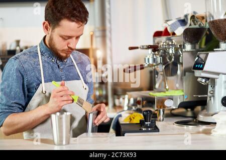 Junger bärtiger Mann in Schürze steht an der Theke, hält Pinsel, reinigt Metallteile der Kaffeemaschine, bereitet Platz für die Arbeit, Morgenzeitkonzept Stockfoto