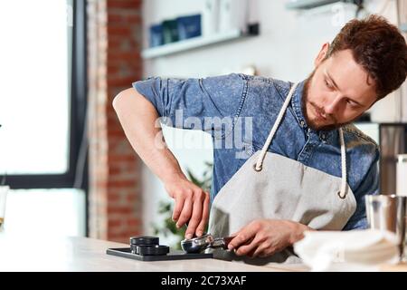 Professioneller junger Mann mit dicken stilvollen Bart, trägt blaue Jeans-Hemd und ordentlich Schürze, drückt Kaffee mit Tamper, schaut auf Kunden, arbeiten Stockfoto