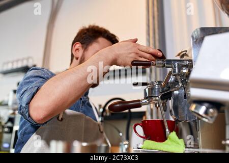 Junge bärtige Mann steht in der Nähe von professionellen Mühle, hält Griff, beobachtet Kaffee Gießen Prozess, bereitet frische aromatische Cappuccino für Kunden in m Stockfoto