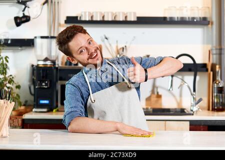 Fröhlich. Optimistischer Mann mit dickem Bart, trägt Jeans-Hemd und gepflegte Schürze, showa Daumen nach oben Geste, gradfully lächelt, schaut auf die Kamera mit glücklichem Gesicht Stockfoto