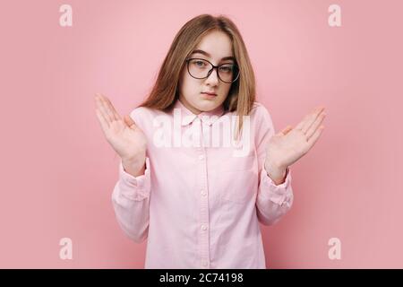 Portrait von frustrierten jungen Mädchen zuckte mit Schultern, um menschliche Emotionen vor der Kamera auszudrücken. Blonde Frau steht über rosa Hintergrund in Brillen Stockfoto