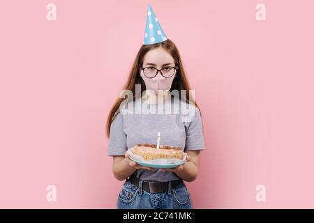 Vorderansicht der jungen Frau mit festlicher Mütze und medizinischer Maske, stehend über rosa Hintergrund und hält Geburtstagskuchen mit einer brennenden Kerze. Konz Stockfoto