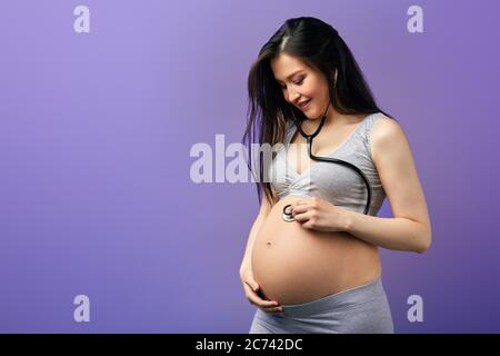 Selbstbewusst lächelnde schwangere Frau in Hauskleidung hören auf ihren Bauch mit einem Stethoskop, Vorbereitung für bald nach Hause Geburt. Geburt, Geburt, Haus B Stockfoto
