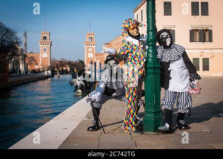 VENEDIG, ITALIEN - 28. FEBRUAR 2020: Harlekin Maske posiert zusammen mit zwei anderen schwarz-weißen Masken. Im Hintergrund das Arsenale von Venedig Stockfoto