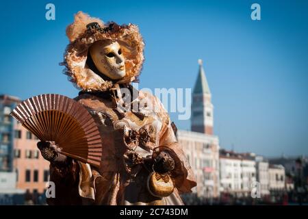 VENEDIG, ITALIEN - 28. FEBRUAR 2020: Eine goldene Maske während des Karnevals in Venedig winkt einem Fächer mit dem Markusplatz im Hintergrund Stockfoto