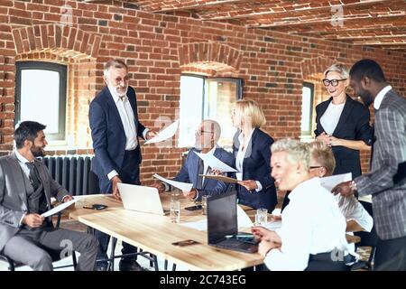 Gruppe von Geschäftsleuten tragen stilvolle Anzüge am Tisch Projekt diskutieren. Kaukasisch Erwachsene blonde Mann tragen blauen Anzug stehen, sprechen. Andere hören Sie genau zu Stockfoto