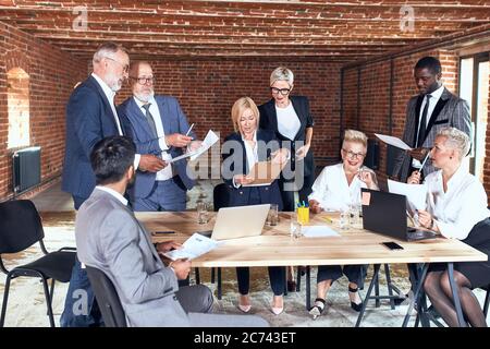 Gruppe von acht Geschäftsleuten tragen Büroanzüge Brainstorming zusammen, Ideen austauschen, sprechen im Besprechungsraum mit Backsteinwänden. In der Mitte Tabelle auf wh Stockfoto