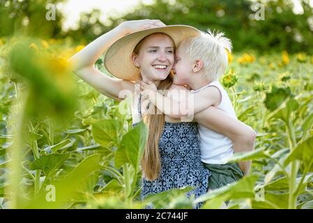 Glücklich lachende Mutter geben Kleinkind Sohn Huckepack Fahrt auf dem Hintergrund der grünen blühenden Sonnenblumen Feld. Stockfoto