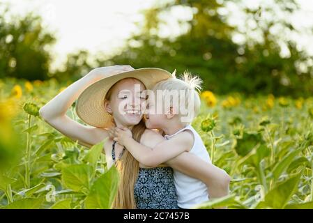 Glücklich lachende Mutter geben Kleinkind Sohn Huckepack Fahrt auf dem Hintergrund der grünen blühenden Sonnenblumen Feld. Stockfoto