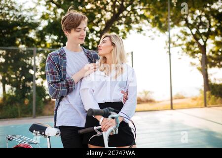 Schönes Mädchen mit blonden Haaren und Junge träumerisch auf einander beim Stehen im Park. Nettes junges Paar umarmt sich gegenseitig mit Fahrrad Stockfoto