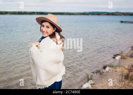 Porträt einer jungen glücklichen Frau, die am Wasser entlang geht und die Seenlandschaft genießt. Reisende mit Decke bedeckt trägt Hut Stockfoto