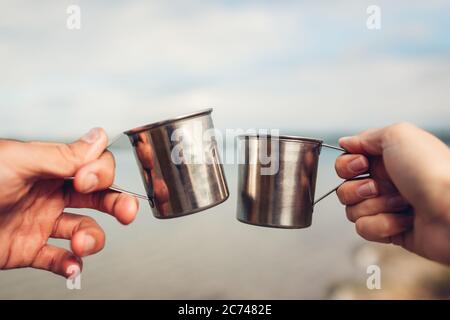 Ein paar Reisende halten heißen Tee in Metallbechern klirren. Menschen trinken warme Getränke am See Stockfoto