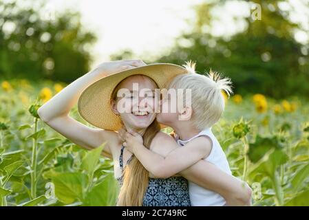 Glücklich lachende Mutter geben Kleinkind Sohn Huckepack Fahrt auf dem Hintergrund der grünen blühenden Sonnenblumen Feld. Stockfoto