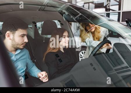 Paar im Auto. Eine Beraterin zeigt einem jungen Paar ein Auto. Junge Familie wählt Auto bei einem Autohaus Stockfoto