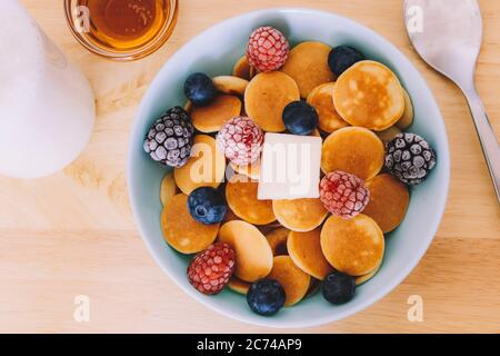 Draufsicht auf eine blaue Schüssel mit Cerealien-Pfannkuchen mit Beeren und Butter neben etwas Sirup auf einem Holztisch Stockfoto